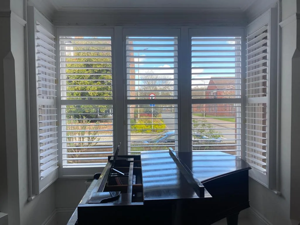 White plantation shutters installed in a bay window in a living room, with a piano in front.
