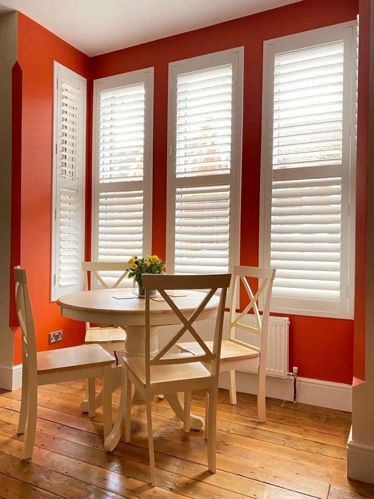 Bright dining room with white full height shutters against a bold orange wall.
