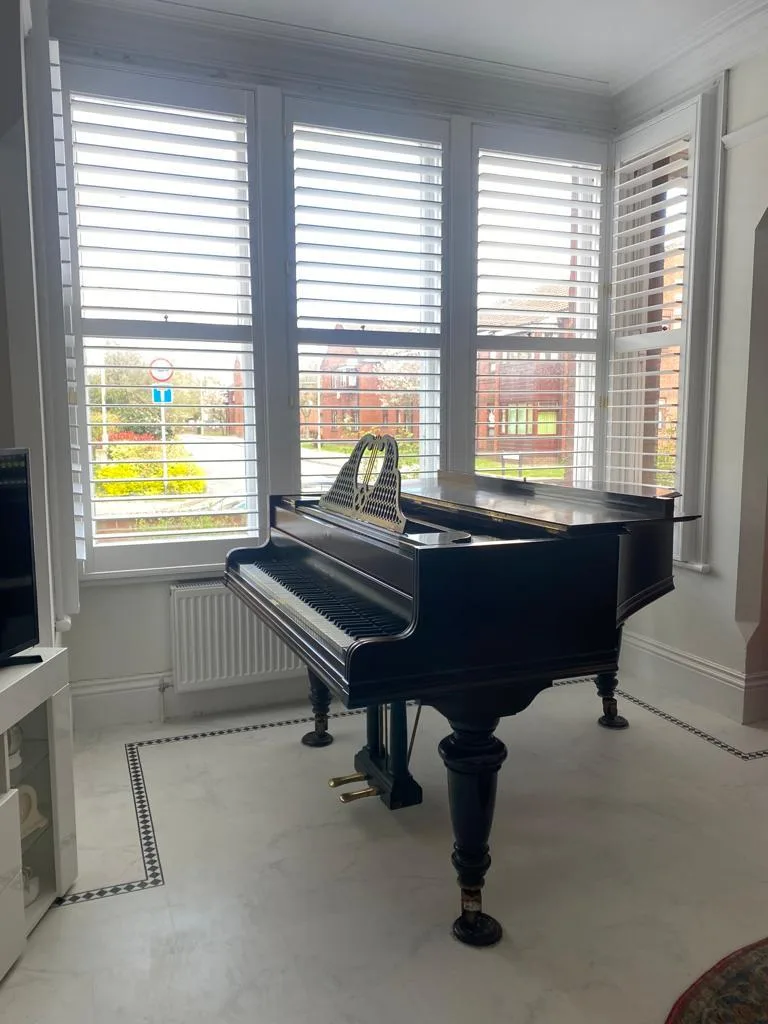 White full height plantation shutters installed in a living room bay window with a grand piano.