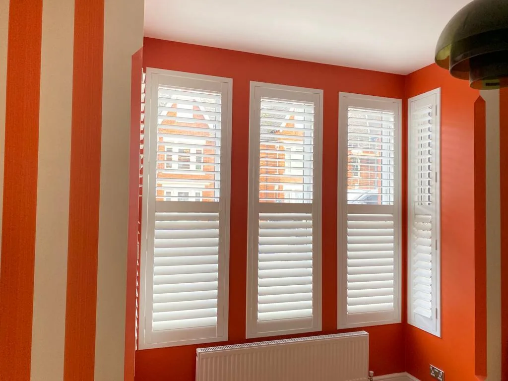 Bright living room with three full height white plantation shutters installed on a coral accent wall.