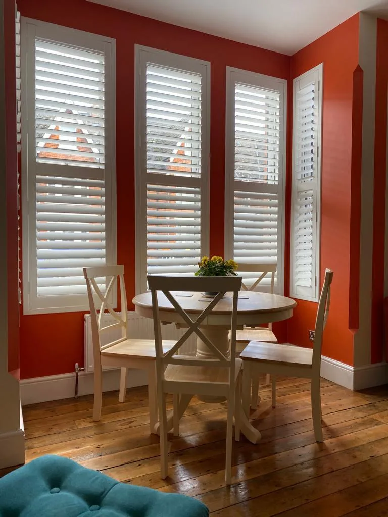 Full height plantation shutters in a dining room with orange walls and wooden floors.
