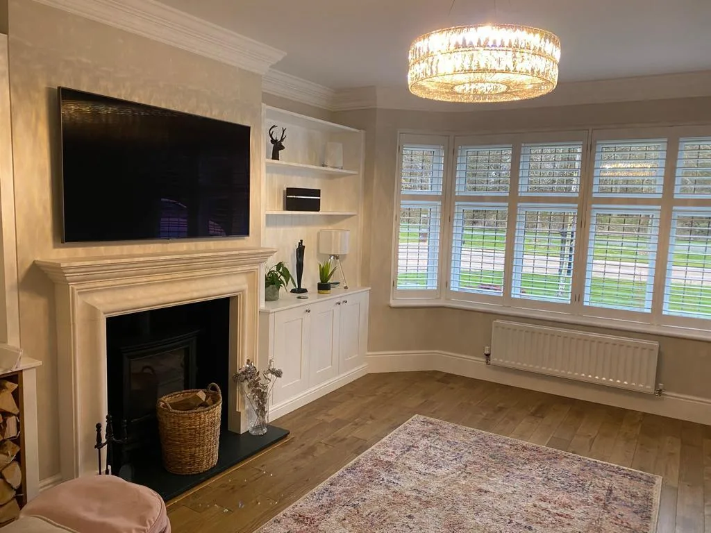 White full height bay window shutters in a living room with a modern fireplace and chandelier.