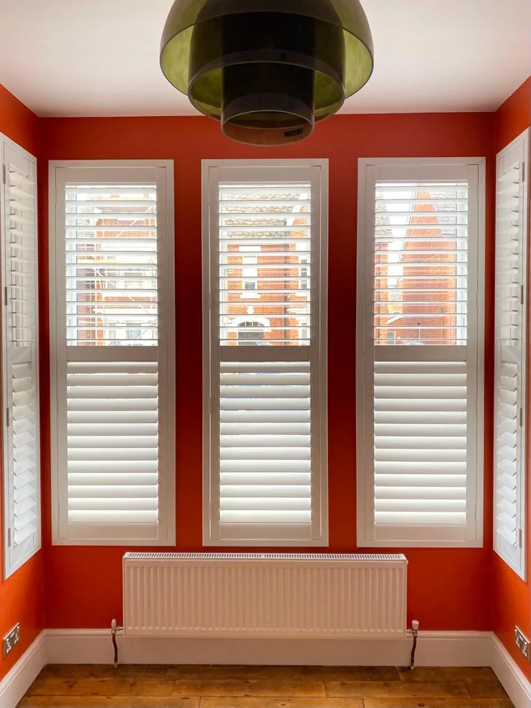 Full height white plantation shutters installed on multiple windows in a room with orange walls.
