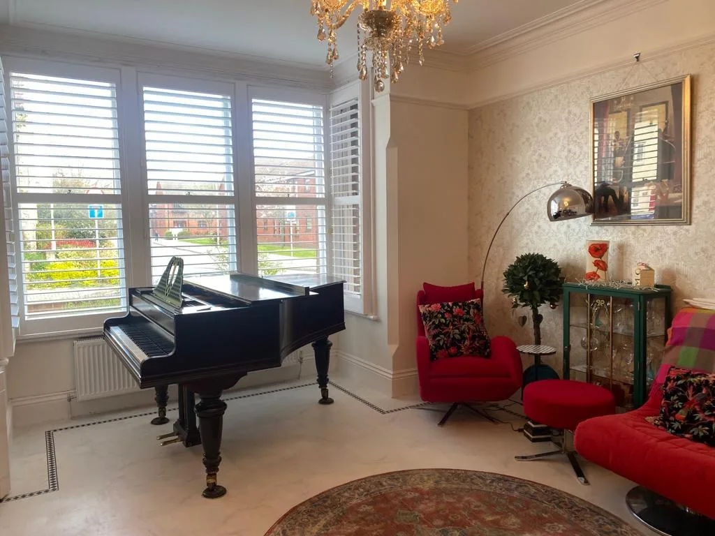 White plantation shutters installed on a bay window in a stylish living room with a piano and red armchairs.
